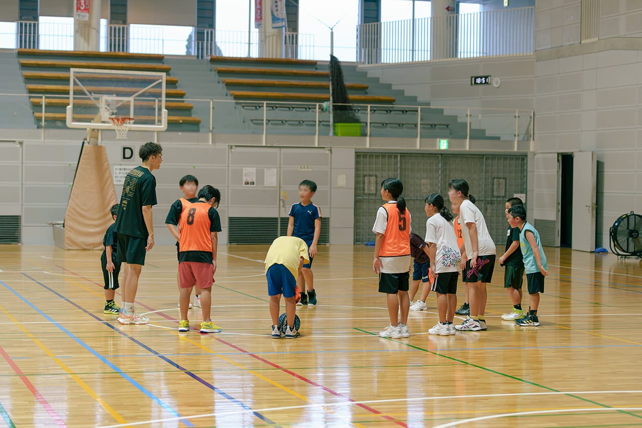 コーチと生徒たちが体育館で円に並びコーチの話を聞いている様子